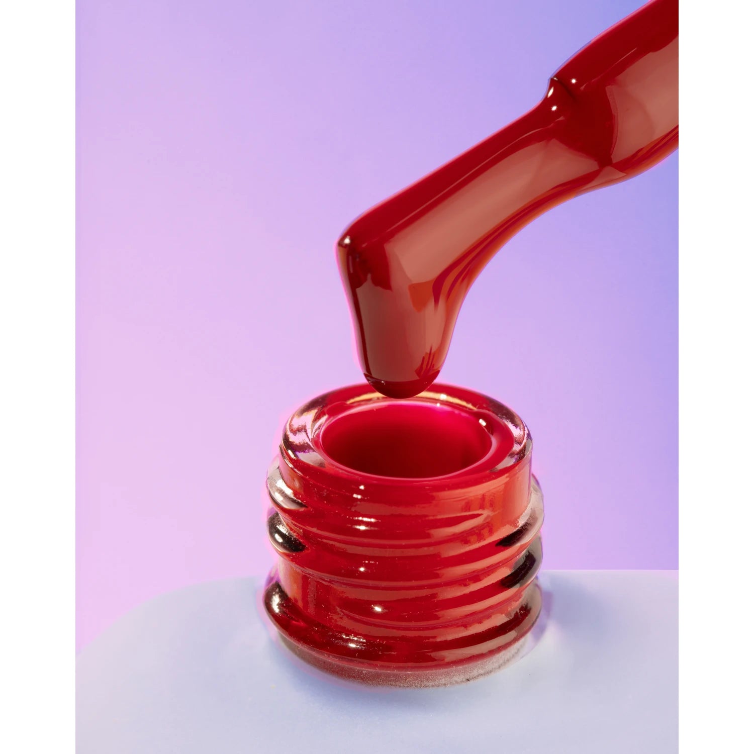 Red gel nail polish being applied with brush, close-up on bottle with purple background