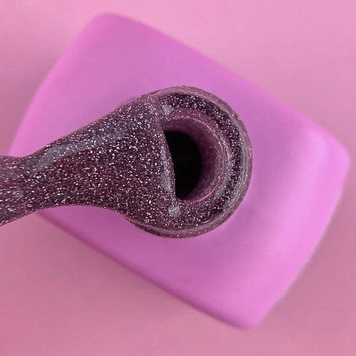 Close-up of glittery purple gel nail polish on brush over pink bottle, manicure product