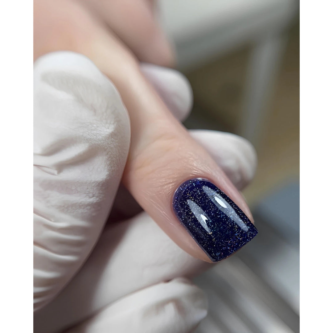 A close-up of a fingernail painted with a deep navy blue glitter nail polish, held by a hand wearing a white glove.