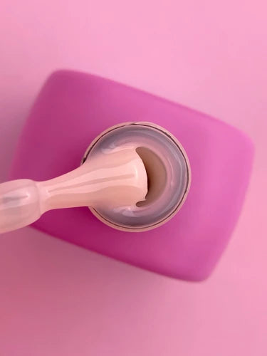 Pale pink UV gel nail polish on brush above open bottle, pink background