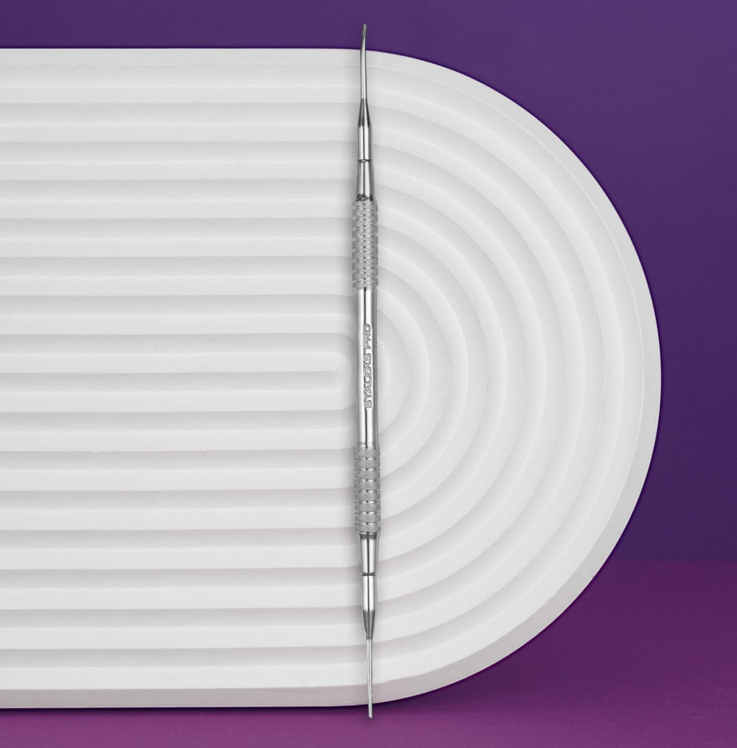Double-ended stainless steel manicure cuticle pusher tool on a white textured surface, purple background.