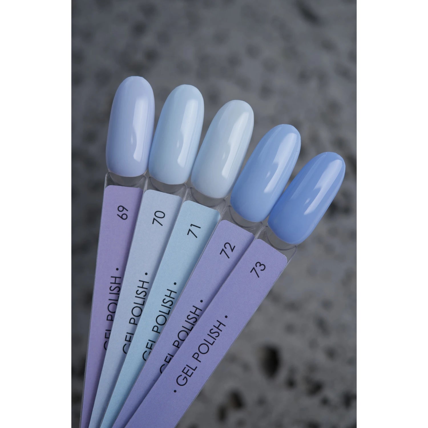 Five blue gel polish nail color samples on display sticks, labeled 69 to 73, on a gray surface.