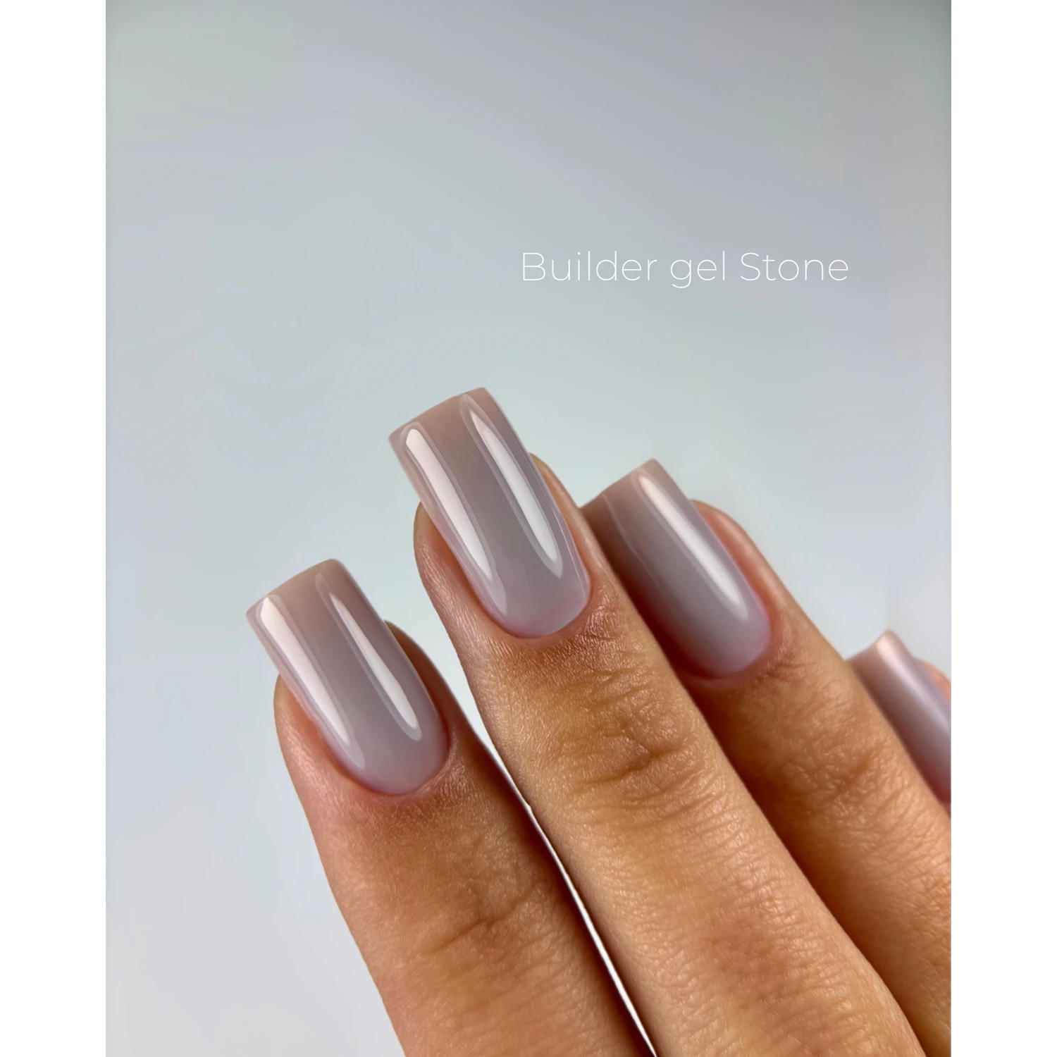 A close-up of a hand with long, square-shaped nails painted in a glossy, pale dusty lavender-gray gel polish, with the text 'Builder gel Stone' printed on the background.