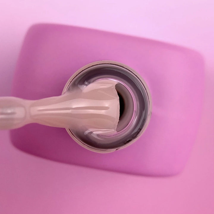 A close-up of a clear nail brush applying a glossy nude-beige nail polish to a circular sample pad on a pink square surface.