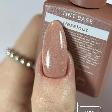 Close-up of a hand with almond-shaped nails painted in shimmery hazelnut gel polish, holding a bottle labeled Tint Base Hazelnut.