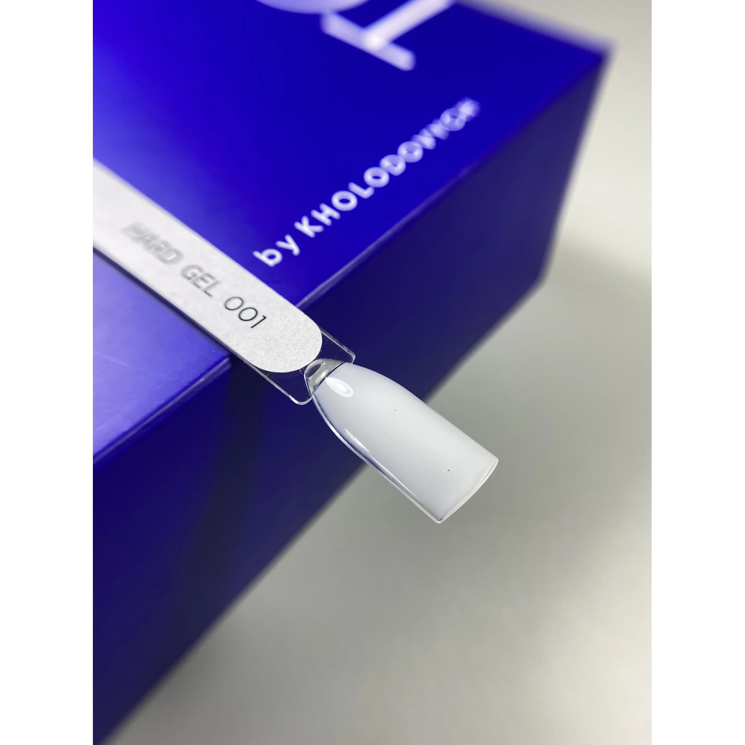 White hard gel nail polish swatch labeled 001 on a display stick next to a blue box