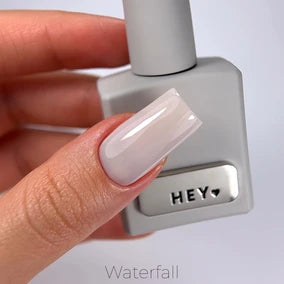 Hand with long glossy nude manicure holding a gray UV gel nail polish bottle labeled HEY.