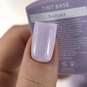 Hand with lilac gel polish manicure, holding a bottle labeled TINT BASE Sunset nail product