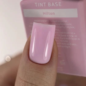 Close-up of a hand with glossy light pink gel nail polish, nail polish bottle labeled 'Tint Base Hilton' in background.