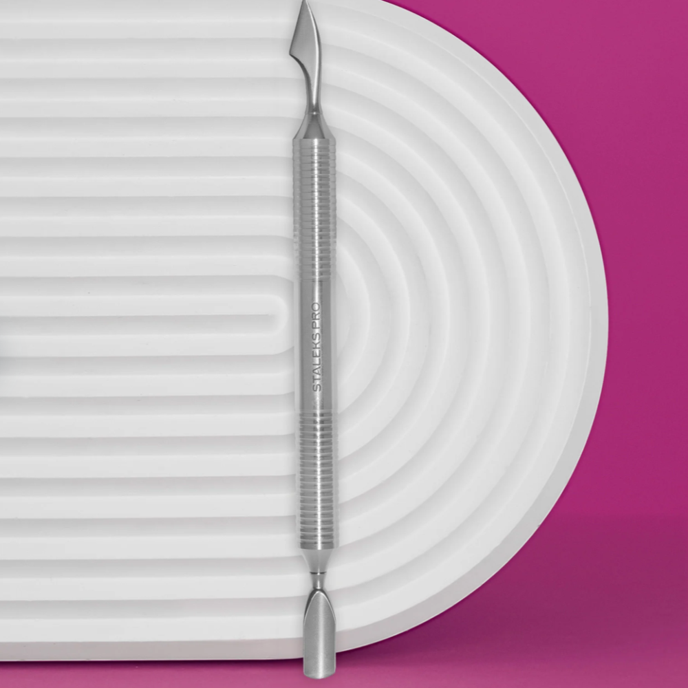 Stainless steel dual-ended nail cuticle pusher on white textured mat with pink background