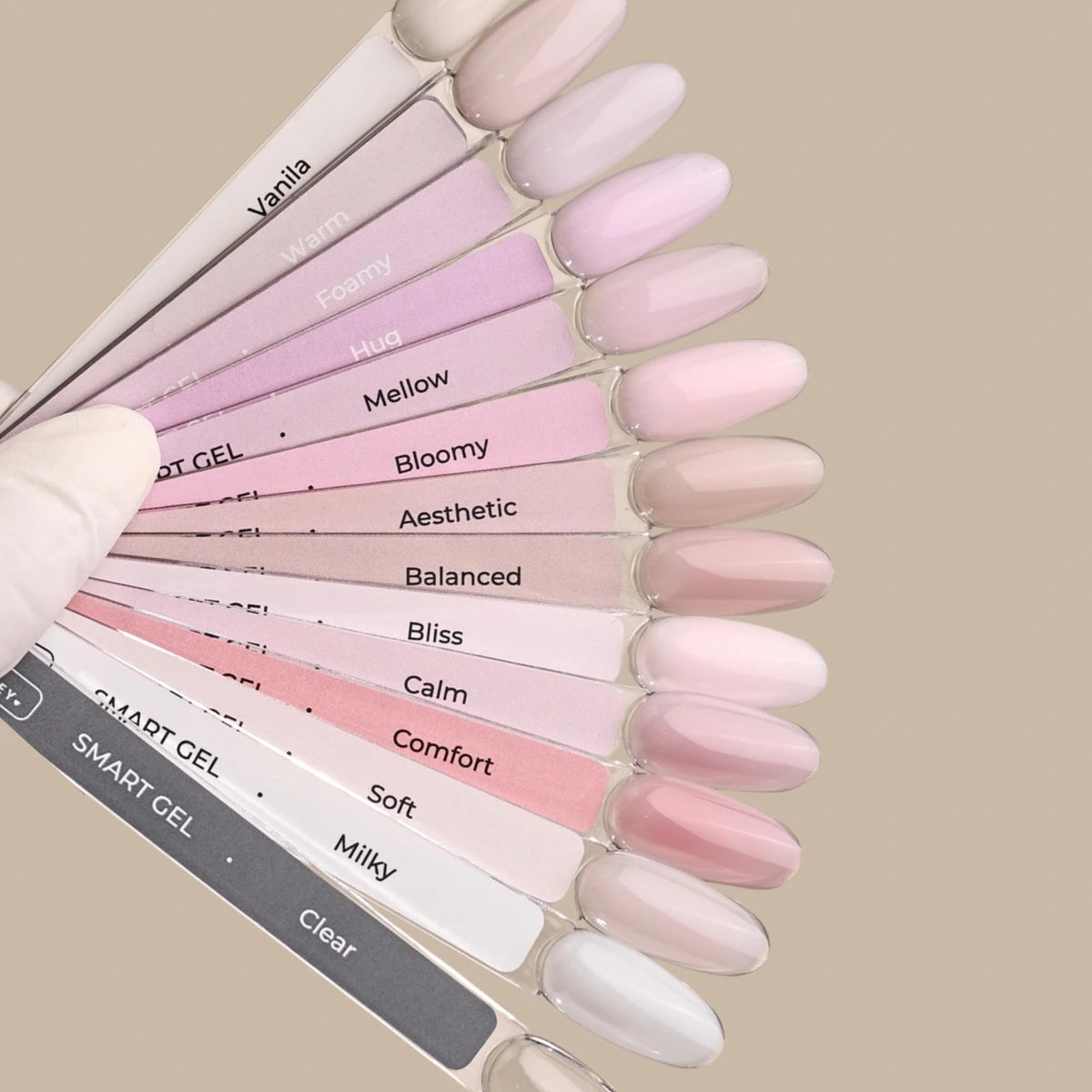 A hand wearing a white glove holds a fan of nail polish sample sticks labeled with names like Vanilla, Foamy, Hug, Mellow, Bloomy, Aesthetic, Balanced, Bliss, Calm, Comfort, soft, Milky, and Clear on a warm beige background.