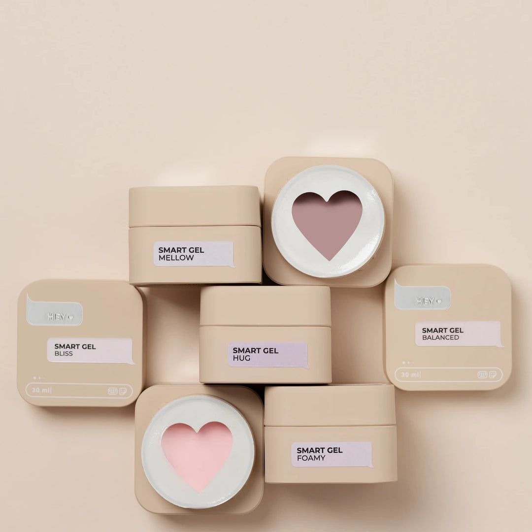 Six beige cosmetic jars arranged in a cluster on a warm beige background. Five jars are closed with square lids, and one open jar at the center shows a heart-shaped swatch of pink gel makeup.