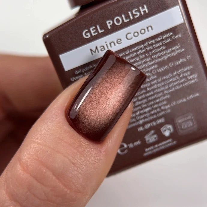 Glossy brown cat eye gel polish manicure on fingernail, bottle labeled 'Maine Coon' in background