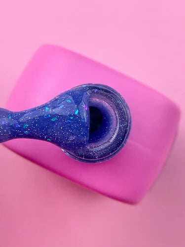 Purple glitter gel nail polish being applied, in a pink bottle, on a pink background.