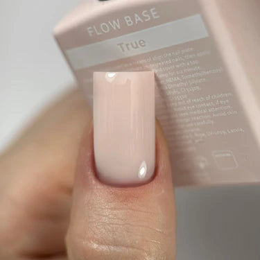 Close-up of a hand with a glossy nude pink gel manicure holding a bottle labeled 'FLOW BASE True'.