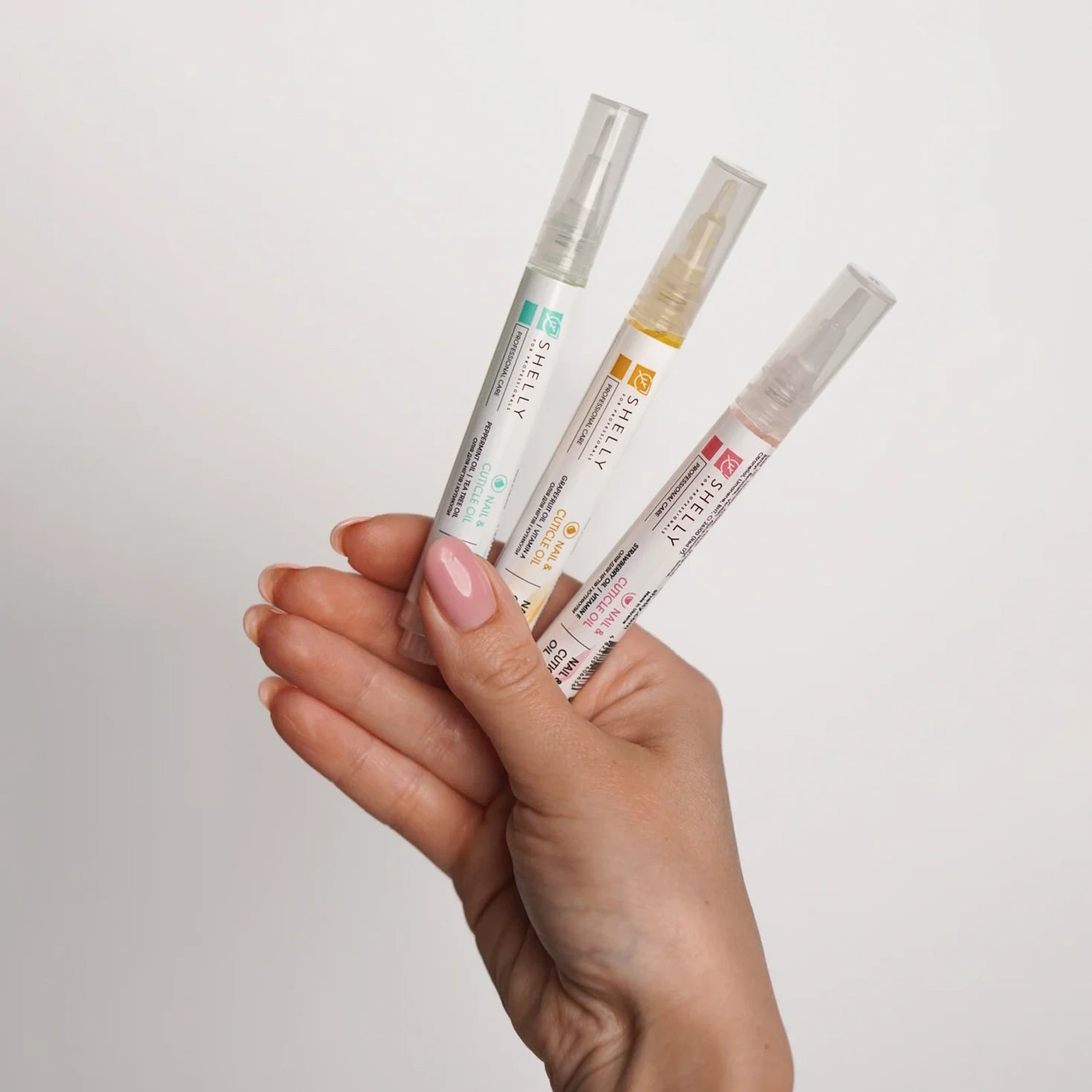 Hand holding three nail care oil pens for cuticle and nail treatment against white background