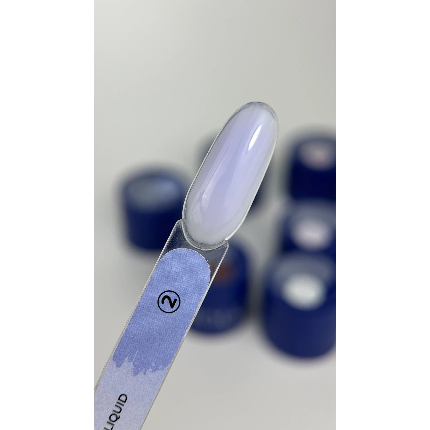 Swatch of sheer light purple UV gel nail polish on sample stick, blue manicure jars in background