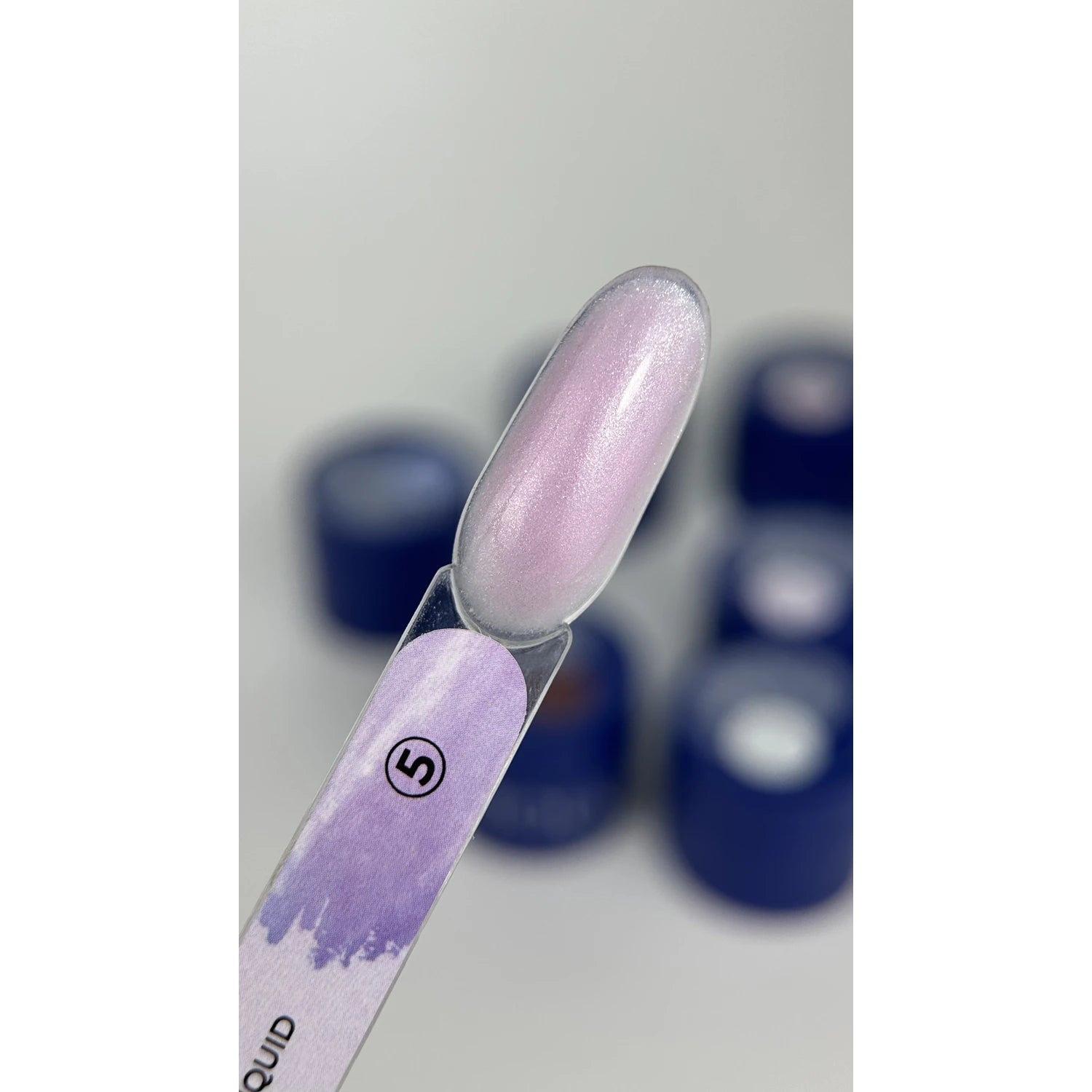 Shimmery pale pink gel polish swatch on nail display stick with blue nail product jars in background