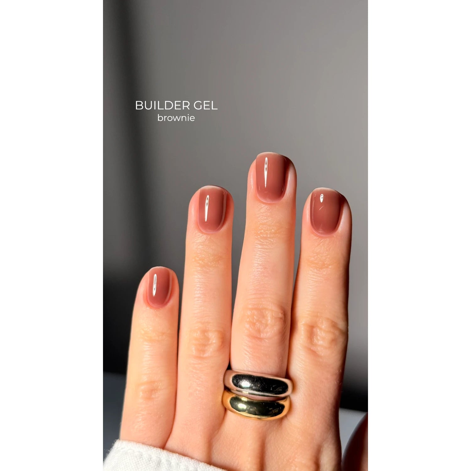 Hand with short nails polished in glossy brown builder gel, wearing two metallic rings.