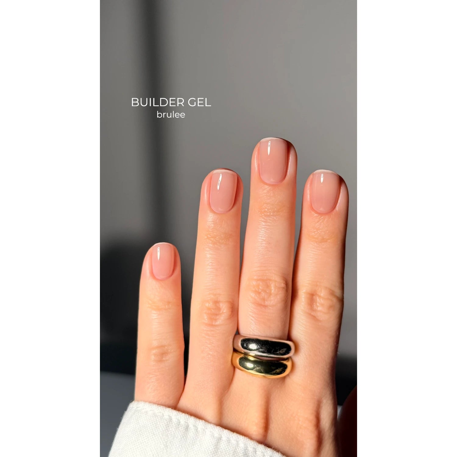 Hand with glossy nude pink builder gel manicure, short nails, and two metallic rings