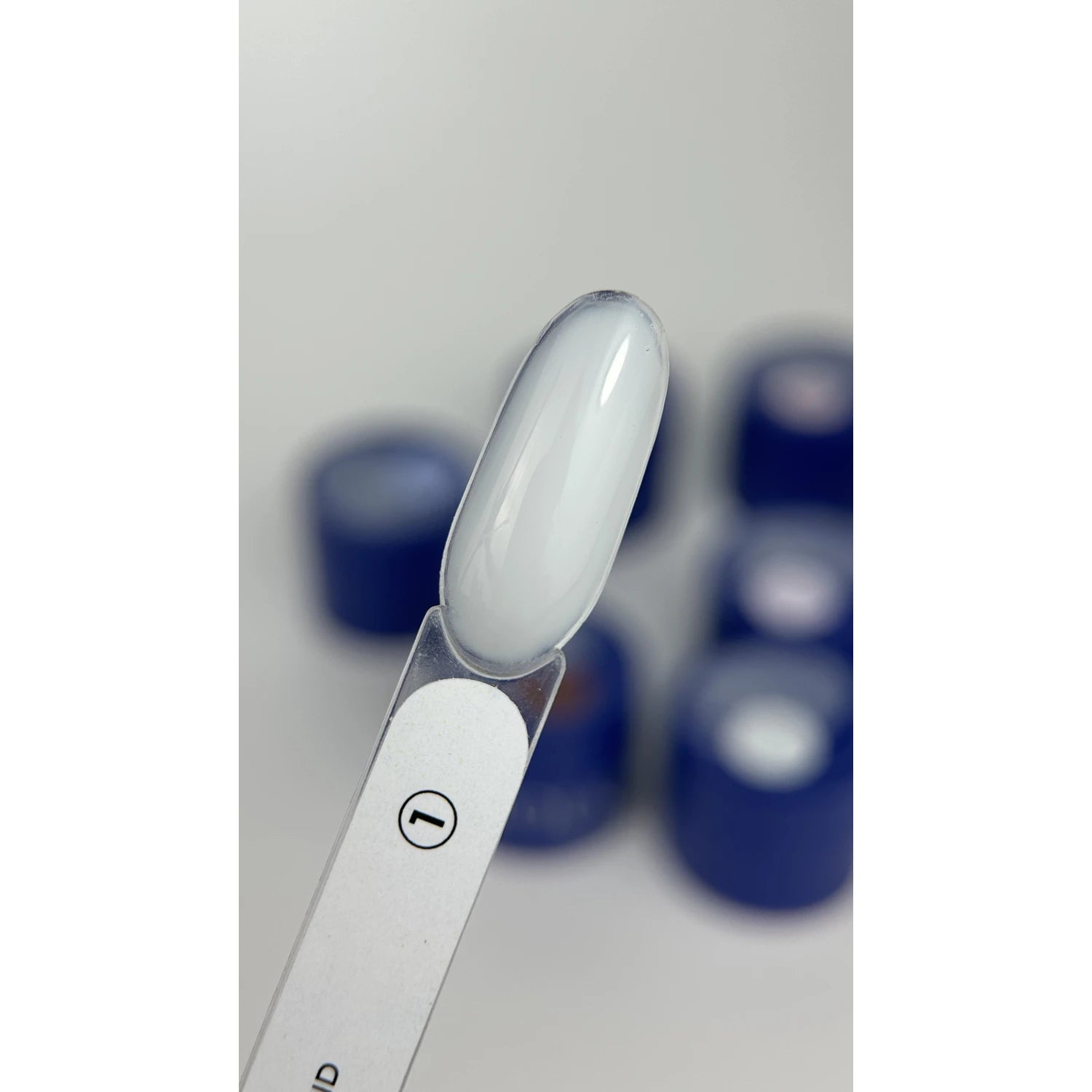 Swatch of milky white UV gel nail polish on sample stick with blue polish jars in background