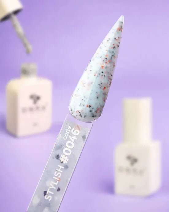 Swatch of speckled blue gel nail polish on sample stick, code #0046, with bottles in background