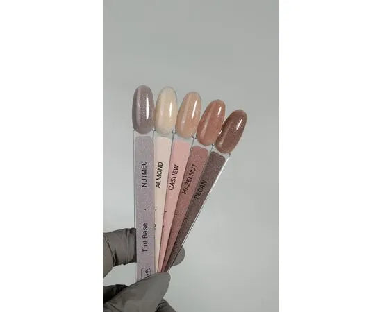 Hand holding nail polish swatches in neutral shades, labeled Nutmeg, Almond, Cashew, Hazelnut, Pecan.