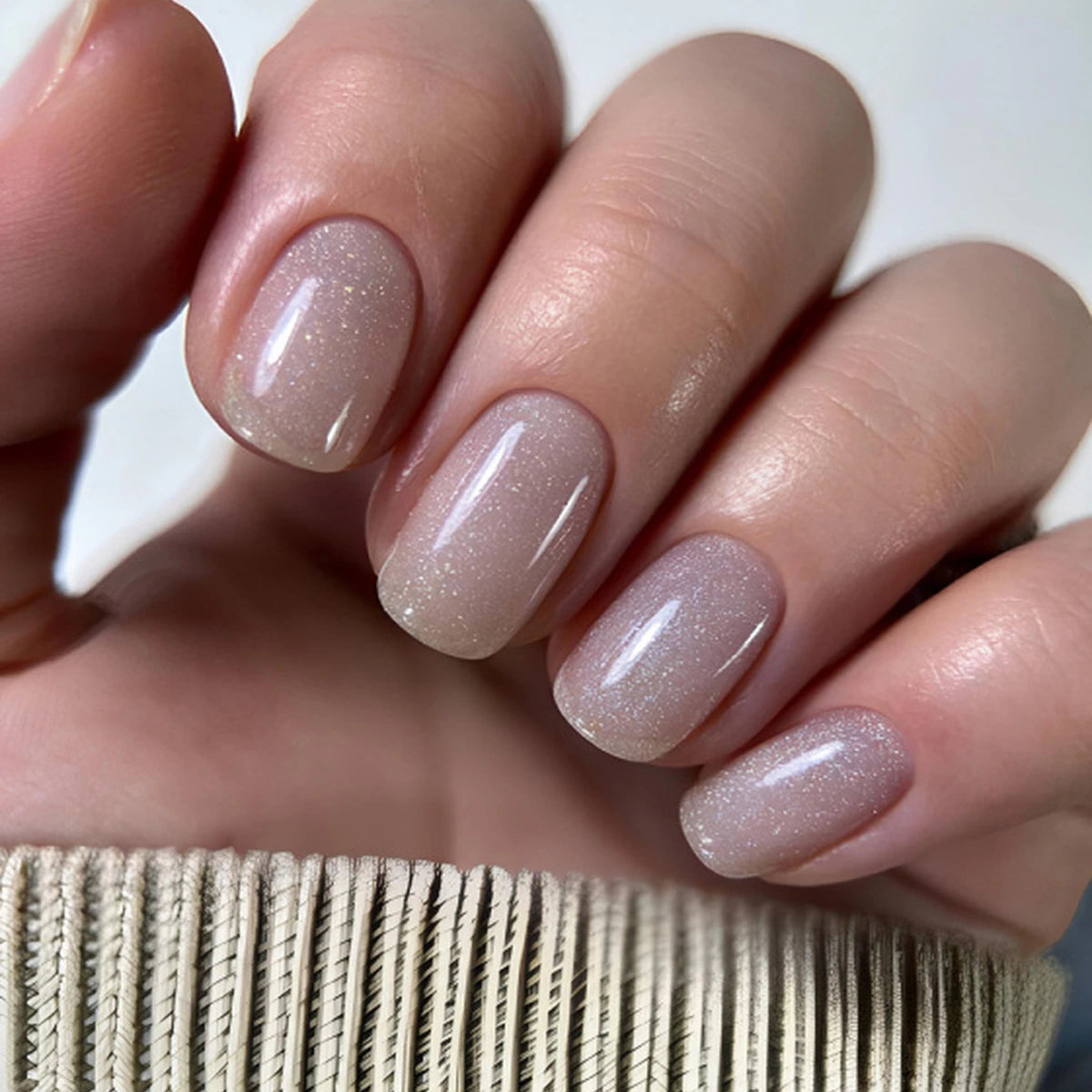 Close-up of a hand with manicured nails painted in a glossy, sheer nude-beige polish with fine shimmer, resting above a ribbed beige knit sleeve.