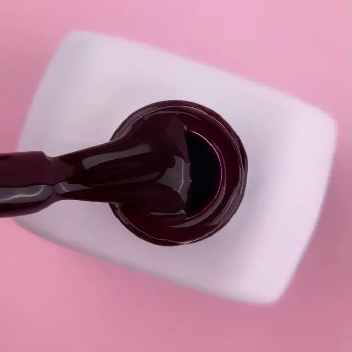 Dark burgundy gel nail polish bottle with brush on a pink background