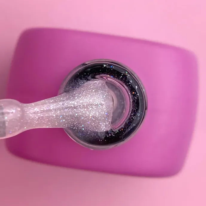 Open nail polish bottle with glittery gel polish on brush, pink background